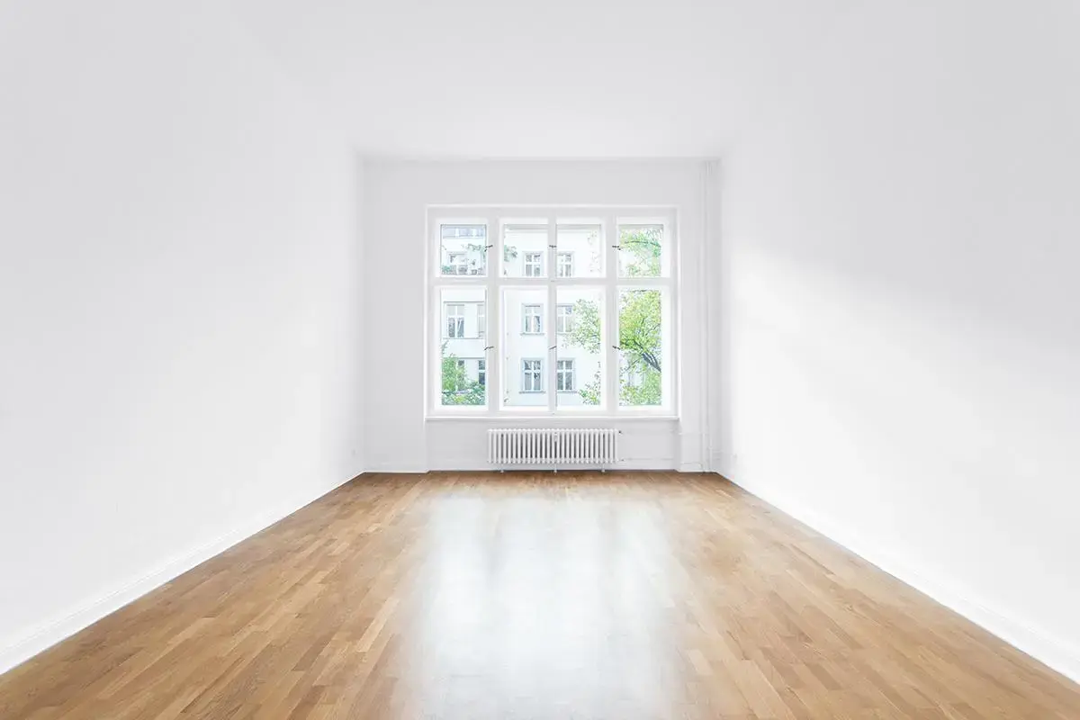 Pièce vide aux murs blancs avec un parquet clair et une grande fenêtre donnant sur des arbres et un immeuble.